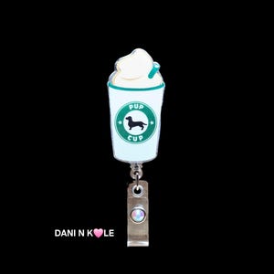 May include: A white and green coffee cup with a black dachshund silhouette and the text "Pup Cup" on the side. The cup has a white whipped cream topping and a green straw. The cup is attached to a silver retractable badge holder with a clear rhinestone.
