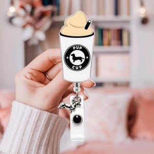 May include: A badge reel featuring a white cup graphic with a black circular logo containing a silhouette of a dachshund and the text PUP CUP. The cup is topped with tan whipped cream and a black straw. A small silver dachshund charm hangs below.