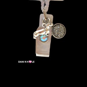 May include: A badge reel featuring a rectangular top piece with the text THE DACHSHUND MOM and an illustration of orange roses. A silver-toned dachshund charm and a circular charm with a textured, shimmering silver surface hang from the reel.