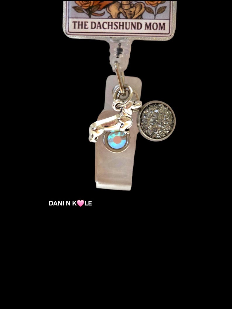 May include: A badge reel featuring a rectangular top piece with the text THE DACHSHUND MOM and an illustration of orange roses. A silver-toned dachshund charm and a circular charm with a textured, shimmering silver surface hang from the reel.