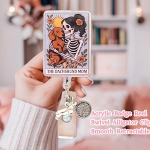May include: An acrylic badge reel featuring a rectangular tarot card design with a skeleton holding a brown dachshund, surrounded by orange roses and a sun. The text reads THE DACHSHUND MOM. It includes a swivel alligator clip and a retractable cord.
