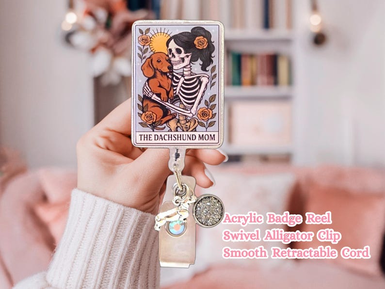 May include: An acrylic badge reel featuring a rectangular tarot card design with a skeleton holding a brown dachshund, surrounded by orange roses and a sun. The text reads THE DACHSHUND MOM. It includes a swivel alligator clip and a retractable cord.