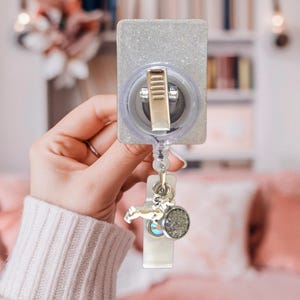 May include: A rectangular badge reel with a silver glitter finish and a metallic clip on the back. A clear plastic strap extends downward, featuring a small silver-toned charm shaped like a dog and a circular pendant with a textured grey surface.