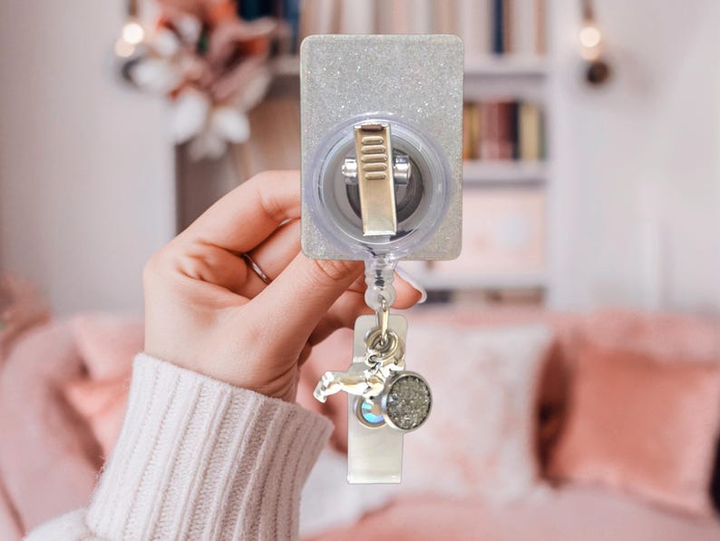 May include: A rectangular badge reel with a silver glitter finish and a metallic clip on the back. A clear plastic strap extends downward, featuring a small silver-toned charm shaped like a dog and a circular pendant with a textured grey surface.