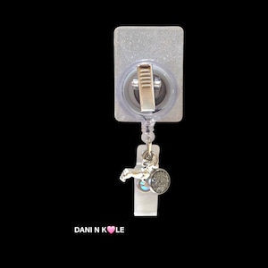 May include: A rectangular badge reel with a glittery finish, featuring a metal clip on the back and a retractable cord. Attached to the cord is a silver-toned charm and a small circular textured pendant. The text DANI N K&hearts;LE is printed below.