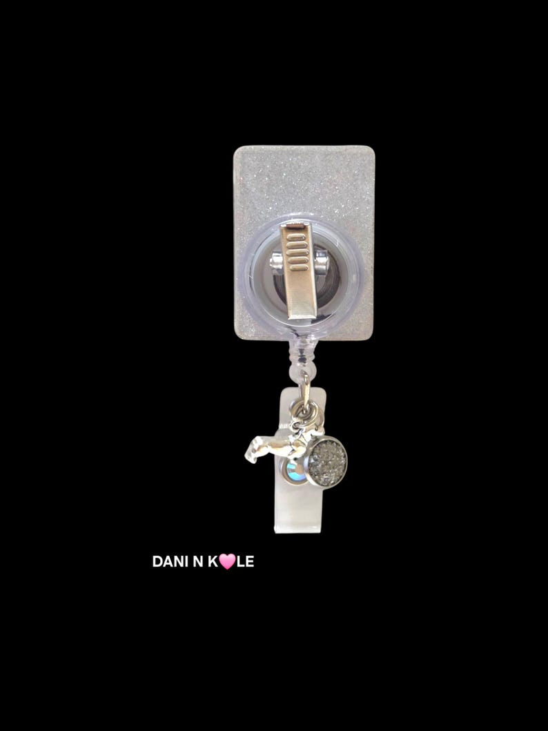 May include: A rectangular badge reel with a glittery finish, featuring a metal clip on the back and a retractable cord. Attached to the cord is a silver-toned charm and a small circular textured pendant. The text DANI N K&hearts;LE is printed below.