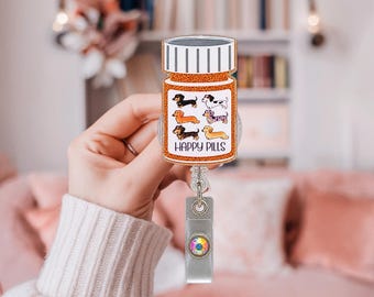 May include: A badge reel featuring a glittery orange pill bottle shape with a white cap. The label displays six illustrated dachshunds in various colors and the text HAPPY PILLS in black. A clear plastic strap with a metal clip hangs below.