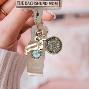 May include: A retractable badge reel featuring a rectangular top plate with the text THE DACHSHUND MOM. Attached to the reel are two charms: a silver-toned dachshund dog silhouette and a circular charm with a textured, shimmering gray surface.