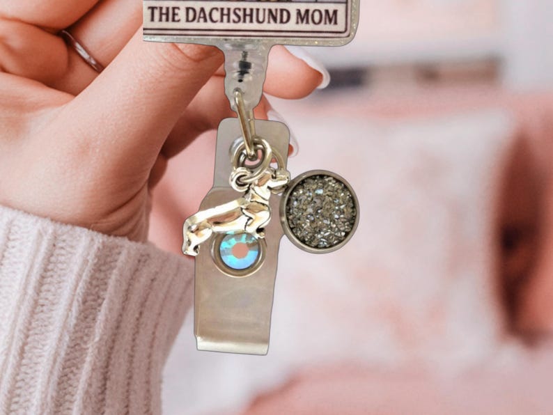 May include: A retractable badge reel featuring a rectangular top plate with the text THE DACHSHUND MOM. Attached to the reel are two charms: a silver-toned dachshund dog silhouette and a circular charm with a textured, shimmering gray surface.