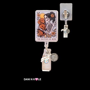 May include: A rectangular badge reel featuring a tarot card illustration of a skeleton holding a brown dachshund, surrounded by orange roses and a sun. The text reads THE DACHSHUND MOM. It includes a silver-toned charm and a circular glittery accent.