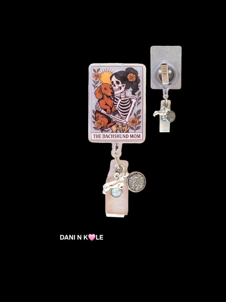 May include: A rectangular badge reel featuring a tarot card illustration of a skeleton holding a brown dachshund, surrounded by orange roses and a sun. The text reads THE DACHSHUND MOM. It includes a silver-toned charm and a circular glittery accent.