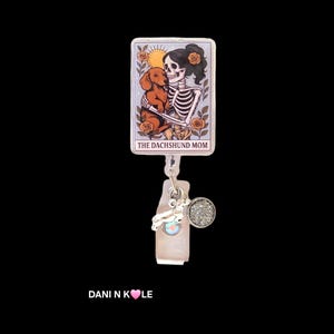 May include: A rectangular badge reel featuring a tarot card illustration of a skeleton holding a brown dachshund, surrounded by orange roses and a sun. The text reads THE DACHSHUND MOM. A silver charm and a small circular glitter accent are attached.