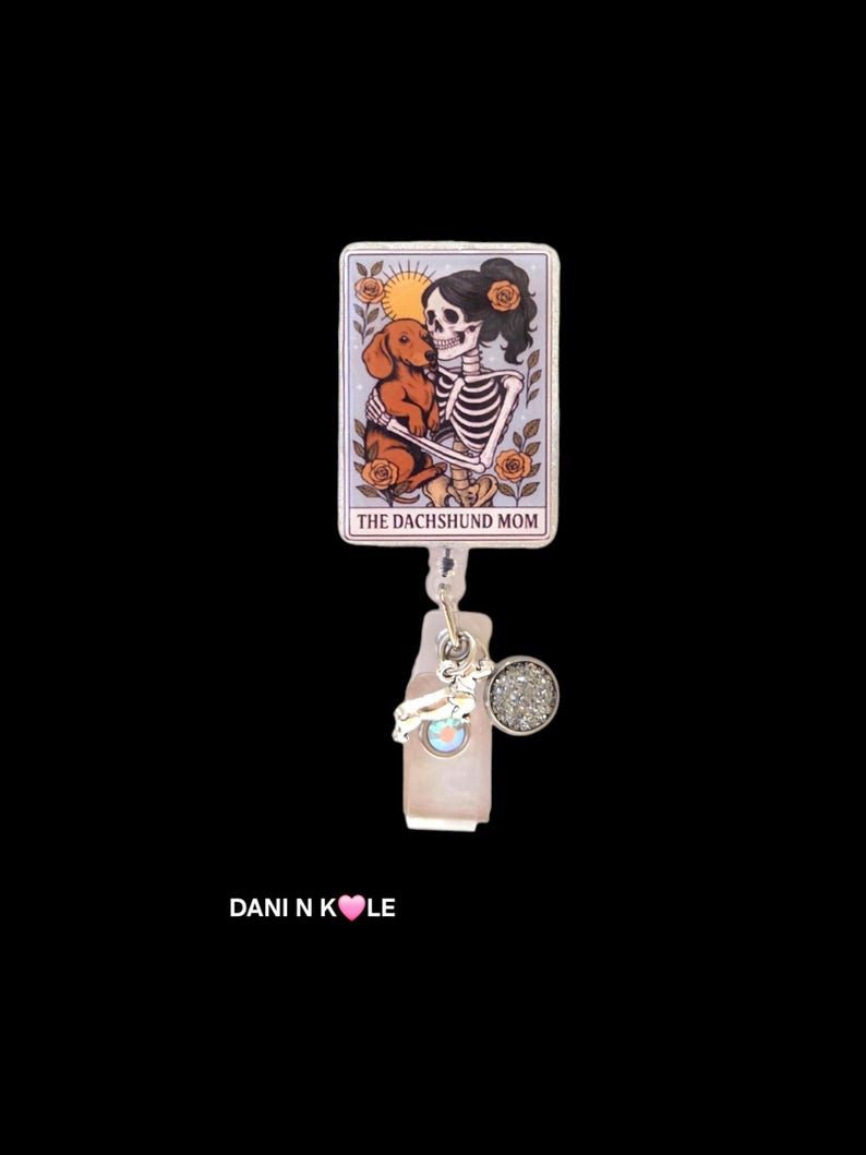 May include: A rectangular badge reel featuring a tarot card illustration of a skeleton holding a brown dachshund, surrounded by orange roses and a sun. The text reads THE DACHSHUND MOM. A silver charm and a small circular glitter accent are attached.