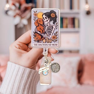 May include: A rectangular badge reel featuring a tarot card illustration of a skeleton holding a brown dachshund, surrounded by orange roses and a sun. The text THE DACHSHUND MOM is printed at the bottom. It includes a silver dachshund charm and a circular glitter charm.