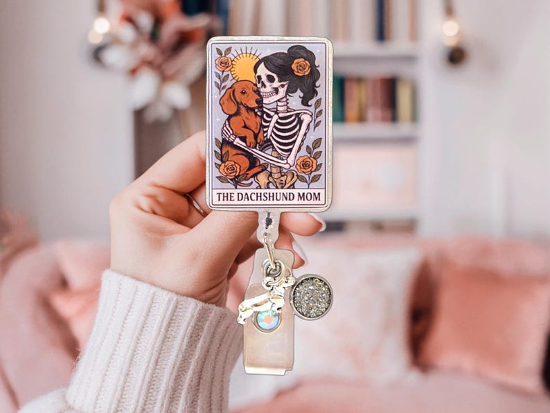 May include: A rectangular badge reel featuring a tarot card illustration of a skeleton holding a brown dachshund, surrounded by orange roses and a sun. The text THE DACHSHUND MOM is printed at the bottom. It includes a silver dachshund charm and a circular glitter charm.