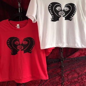 May include: Two t-shirts, one red and one white, each featuring a black graphic of two stylized faces forming a heart shape. The faces have closed eyes and are kissing. The shirts are hanging against a red patterned background.