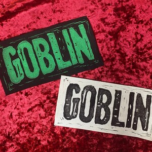 May include: Two fabric patches with the word "GOBLIN" printed on them. The top patch is black with green lettering. The bottom patch is white with black lettering.