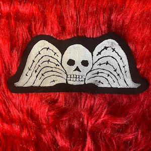 May include: Black and white embroidered patch with a skull and wings. The wings are barbed and the skull has a simple design.