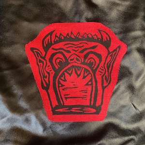 May include: A red patch with a black monster face design is sewn onto a black fabric. The monster has horns, large eyes, and an open mouth with sharp teeth. The patch is a unique, eye-catching design.
