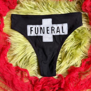 May include: Black ribbed underwear featuring a white cross-shaped patch on the front. The word FUNERAL is printed in black capital letters across the horizontal bar of the white cross patch. The garment has a high-cut leg opening design.