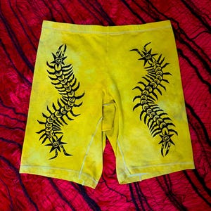 Może przedstawiać: Żółte szorty z czarnym graficznym wzorem przypominającym stonogę na każdej nogawce. Szorty mają efekt tie-dye i wygodny krój. Tło to czerwona tkanina z czarną siatką.