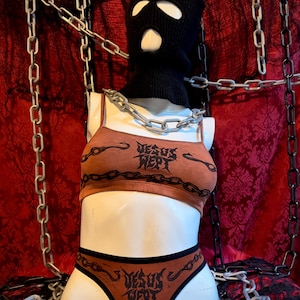 May include: A mannequin head and torso wearing a black balaclava and a brown lingerie set. The lingerie set includes a crop top and underwear, both with the text "JESUS WEPT" in a gothic font. A silver chain is around the neck and chains hang in the background.