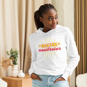 May include: White long-sleeve hooded sweatshirt with the text "MASTER manifestor" in pink and yellow letters with gold stars and a gold starburst design.