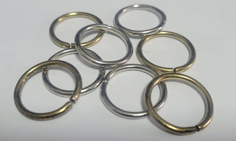 Pu&ograve; includere: Una collezione di anelli aperti in varie finiture metalliche, tra cui argento e oro. Gli anelli sono sparsi su una superficie bianca, mostrando la loro forma circolare e il design aperto, ideali per la creazione di gioielli o lavori artigianali.