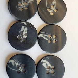 Puede incluir: Seis botones redondos de color gris oscuro con un patrón de remolino blanco veteado. Cada botón tiene cuatro agujeros para coser. Los botones están dispuestos en dos filas de tres sobre un fondo blanco.