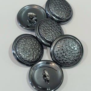 Op de afbeelding: Een verzameling van zeven donkergrijze, ronde knopen. De knopen hebben een getextureerd, kiezelachtig oppervlak aan de voorkant en een lus aan de achterkant om op te naaien. De knopen zijn op een wit oppervlak geplaatst.