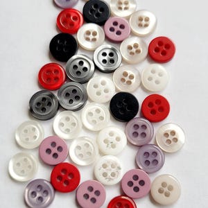 Peut inclure: Un assortiment de boutons de diff&eacute;rentes couleurs, dont rouge, noir, blanc, gris et lavande. Chaque bouton comporte quatre trous et un bord l&eacute;g&egrave;rement sur&eacute;lev&eacute;. Les boutons sont dispos&eacute;s sur une surface blanche, pour la couture et les loisirs cr&eacute;atifs.