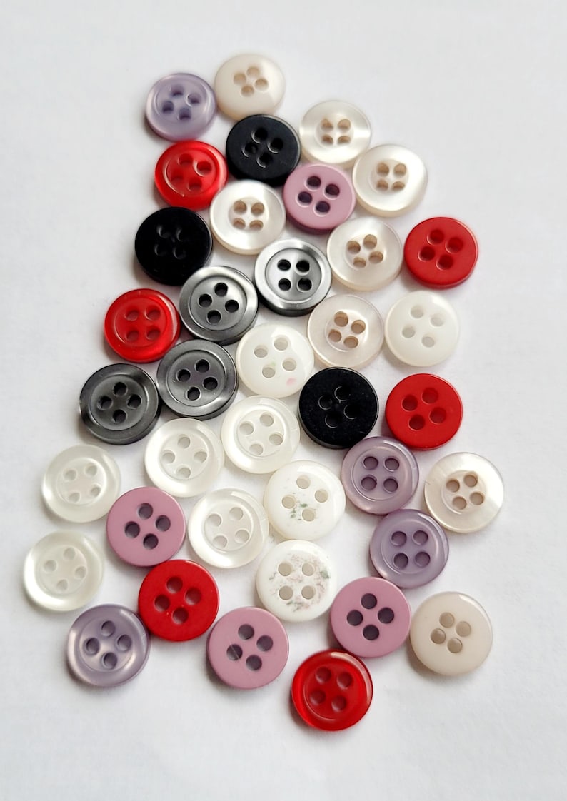 Peut inclure: Un assortiment de boutons de diff&eacute;rentes couleurs, dont rouge, noir, blanc, gris et lavande. Chaque bouton comporte quatre trous et un bord l&eacute;g&egrave;rement sur&eacute;lev&eacute;. Les boutons sont dispos&eacute;s sur une surface blanche, pour la couture et les loisirs cr&eacute;atifs.