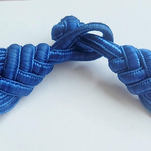 K&ouml;nnte beinhalten: Ein Paar blaue Stoffkn&ouml;pfe oder -kn&ouml;pfe, die miteinander verknotet und gewebt sind, zur Verwendung an Kleidung oder Accessoires.