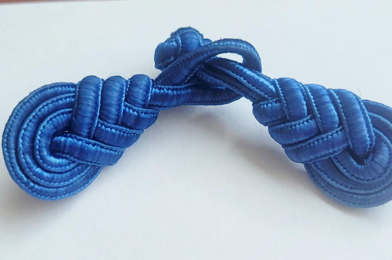 K&ouml;nnte beinhalten: Ein Paar blaue Stoffkn&ouml;pfe oder -kn&ouml;pfe, die miteinander verknotet und gewebt sind, zur Verwendung an Kleidung oder Accessoires.
