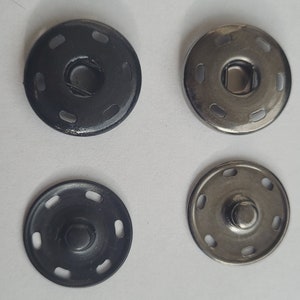 Puede incluir: Cuatro botones a presión de metal, dos negros y dos plateados, con un diseño circular y múltiples agujeros alrededor del borde.