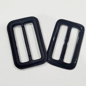 Könnte beinhalten: Zwei schwarze Kunststoff-Schiebeschließen. Die Schließen sind rechteckig mit zwei parallelen Schlitzen in der Mitte.