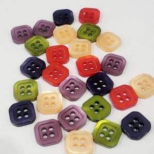 May include: A collection of square, four-hole buttons in a variety of translucent colours including red, purple, green, and light yellow. Each button features rounded corners and a smooth, glossy finish, scattered across a plain white surface.