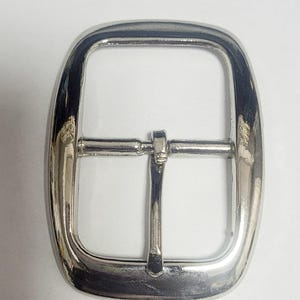 Può includere: Fibbia per cintura argentata lucida, di forma rettangolare con bordi arrotondati. Presenta una barra centrale e un perno per fissare la cintura. Il metallo ha una superficie lucida e riflettente.