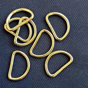 Op de afbeelding: Een groep van zes gouden metalen D-ringen. De ringen liggen los op een donkerblauwe stoffen achtergrond.