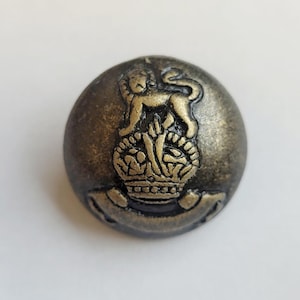 May include: A gold and black metal button featuring a lion standing on a crown with a banner below. The button has a textured surface.