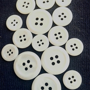 May include: A collection of white, round buttons with four holes each, varying in size. The buttons are arranged on a dark blue textured fabric, creating a contrast between the smooth buttons and the rough background. Ideal for sewing and craft projects.