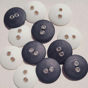 Puede incluir: Una colección de botones redondos en blanco y gris oscuro. Cada botón tiene dos agujeros con anillos metálicos plateados. Los botones están dispuestos en un patrón disperso, adecuados para proyectos de costura o manualidades.