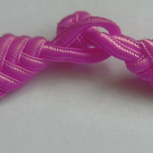 K&ouml;nnte beinhalten: Ein rosa Stoffknopf mit einem geknoteten Design. Der Knopf ist aus einem weichen, seidenartigen Material gefertigt und hat eine gl&auml;nzende Oberfl&auml;che.