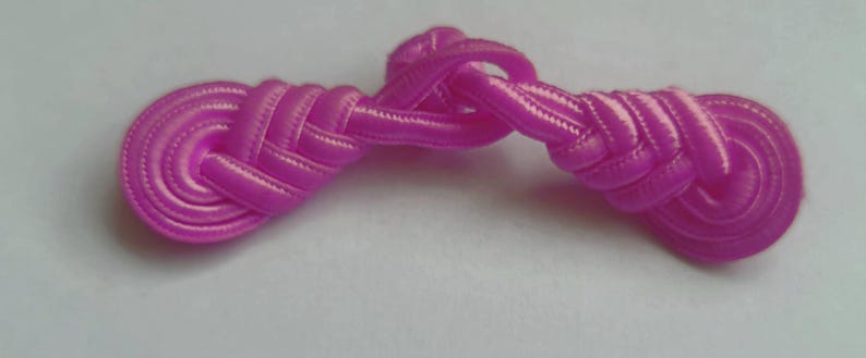 K&ouml;nnte beinhalten: Ein rosa Stoffknopf mit einem geknoteten Design. Der Knopf ist aus einem weichen, seidenartigen Material gefertigt und hat eine gl&auml;nzende Oberfl&auml;che.