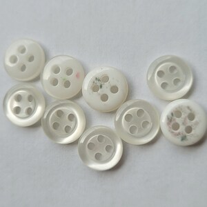 Peut inclure: Collection de neuf boutons ronds blancs &agrave; quatre trous. Les boutons ont un aspect nacr&eacute; et varient l&eacute;g&egrave;rement en taille et en design. Certains pr&eacute;sentent de subtils motifs floraux.