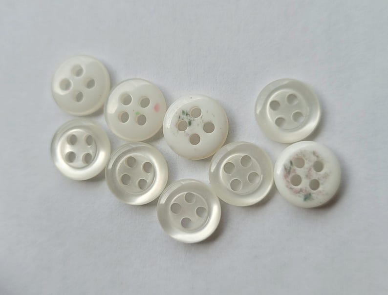 Peut inclure: Collection de neuf boutons ronds blancs &agrave; quatre trous. Les boutons ont un aspect nacr&eacute; et varient l&eacute;g&egrave;rement en taille et en design. Certains pr&eacute;sentent de subtils motifs floraux.