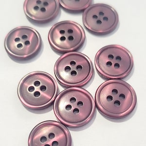 Può includere: Una collezione di dieci bottoni rotondi, dall'aspetto metallico, di colore rosa tenue. Ogni bottone ha quattro fori e un bordo leggermente rialzato. I bottoni sono disposti su una superficie bianca.