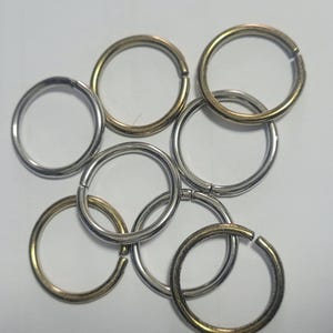 May include: A selection of metal split rings in silver and gold colours. The rings are open, ready for use, and arranged in a random pattern against a white backdrop. These are commonly used for keychains and jewellery making.