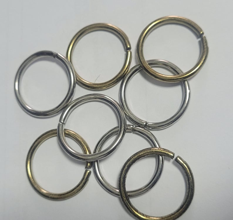 Pu&ograve; includere: Una collezione di anelli spaccati in metallo con finiture varie, tra cui tonalit&agrave; argento e oro. Gli anelli sono aperti, pronti per l'uso, e disposti in modo sparso su uno sfondo bianco. Sono comunemente usati per portachiavi e bigiotteria.