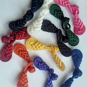K&ouml;nnte beinhalten: Sortiment von 12 bunten, gekn&uuml;pften Stoff-Kn&ouml;pfen oder -Verschl&uuml;ssen. Die Kn&ouml;pfe sind in verschiedenen Farben, darunter Rot, Orange, Gelb, Gr&uuml;n, Blau, Wei&szlig; und Pink.